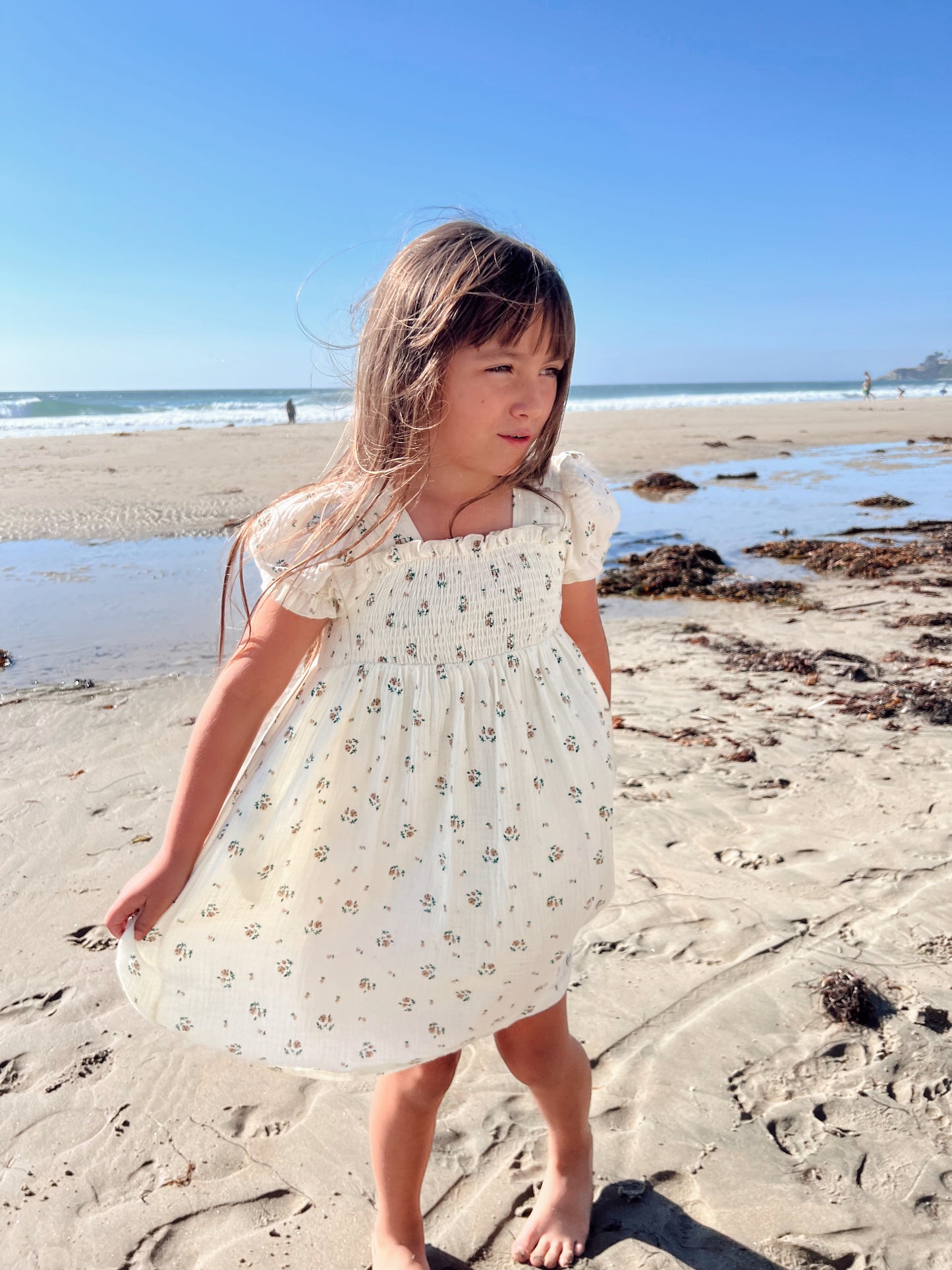 A young girl wearing the "Garden Days" muslin dress from forever french baby stands on a sandy beach with the ocean in the background. She looks off to the side, her hair gently blowing in the breeze. Seaweed is scattered on the shore, and the sky is clear and blue.
