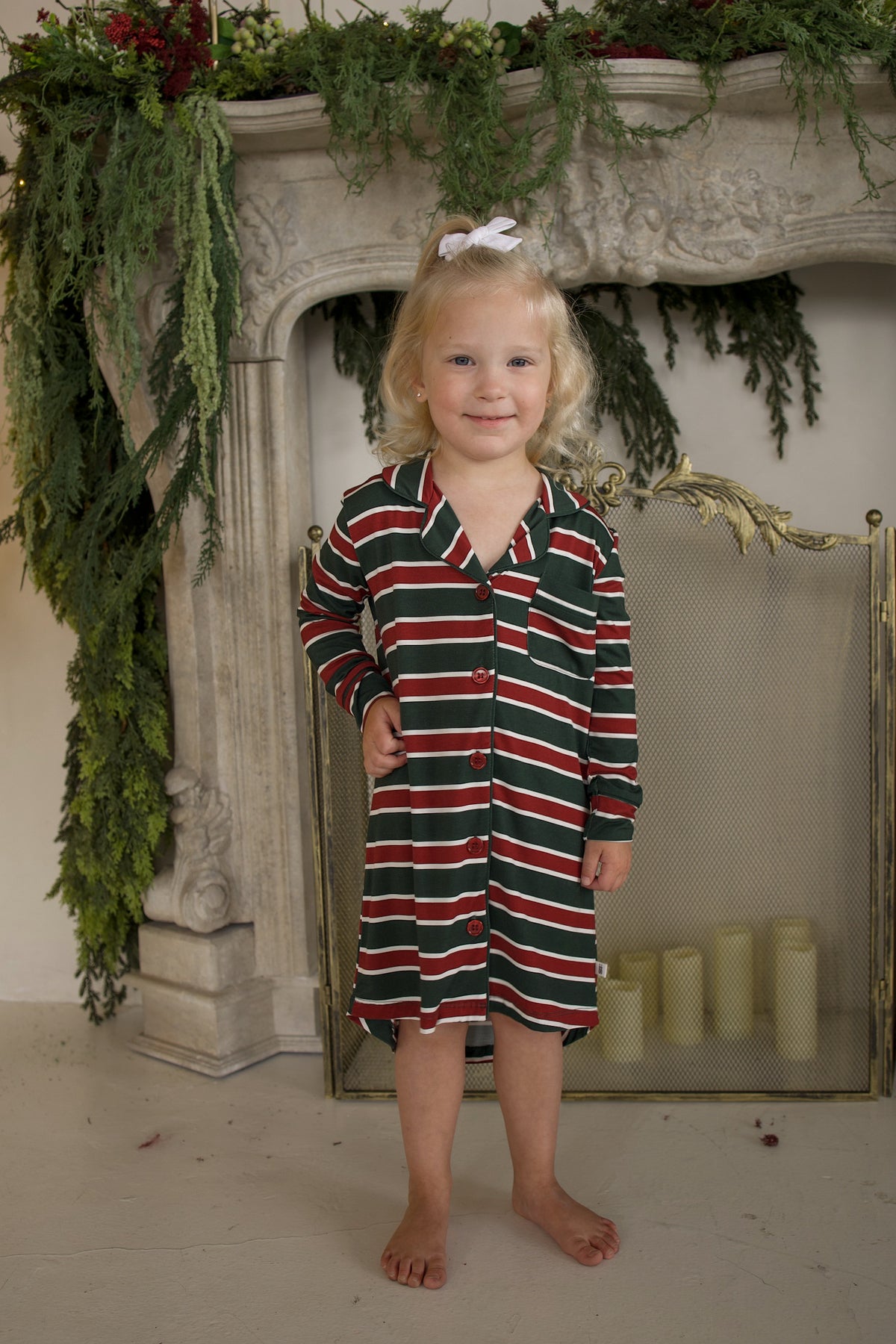 A young girl wearing a Bamboo Sleeping Dress | Holiday Stripe with long sleeves and a button-up front, featuring a red, green, and white holiday stripe pattern.