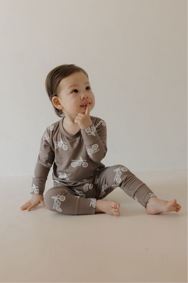A toddler wears the Forever French Baby Bamboo Footless Sleepy Time Set in the Dirt Bike pattern—brown pajamas with a white bicycle design—while sitting on the floor and thoughtfully touching their chin.