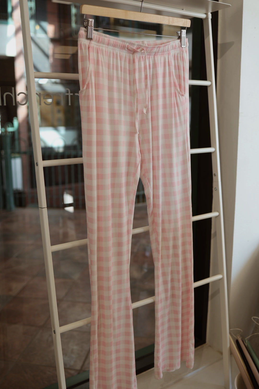 The Long Sleeve Women's Bamboo Pajama in Pink Sugar by forever french baby, shown in light pink and white checks on a hanger by a window, is the perfect comfy and stylish addition to your women’s bamboo lounge set.