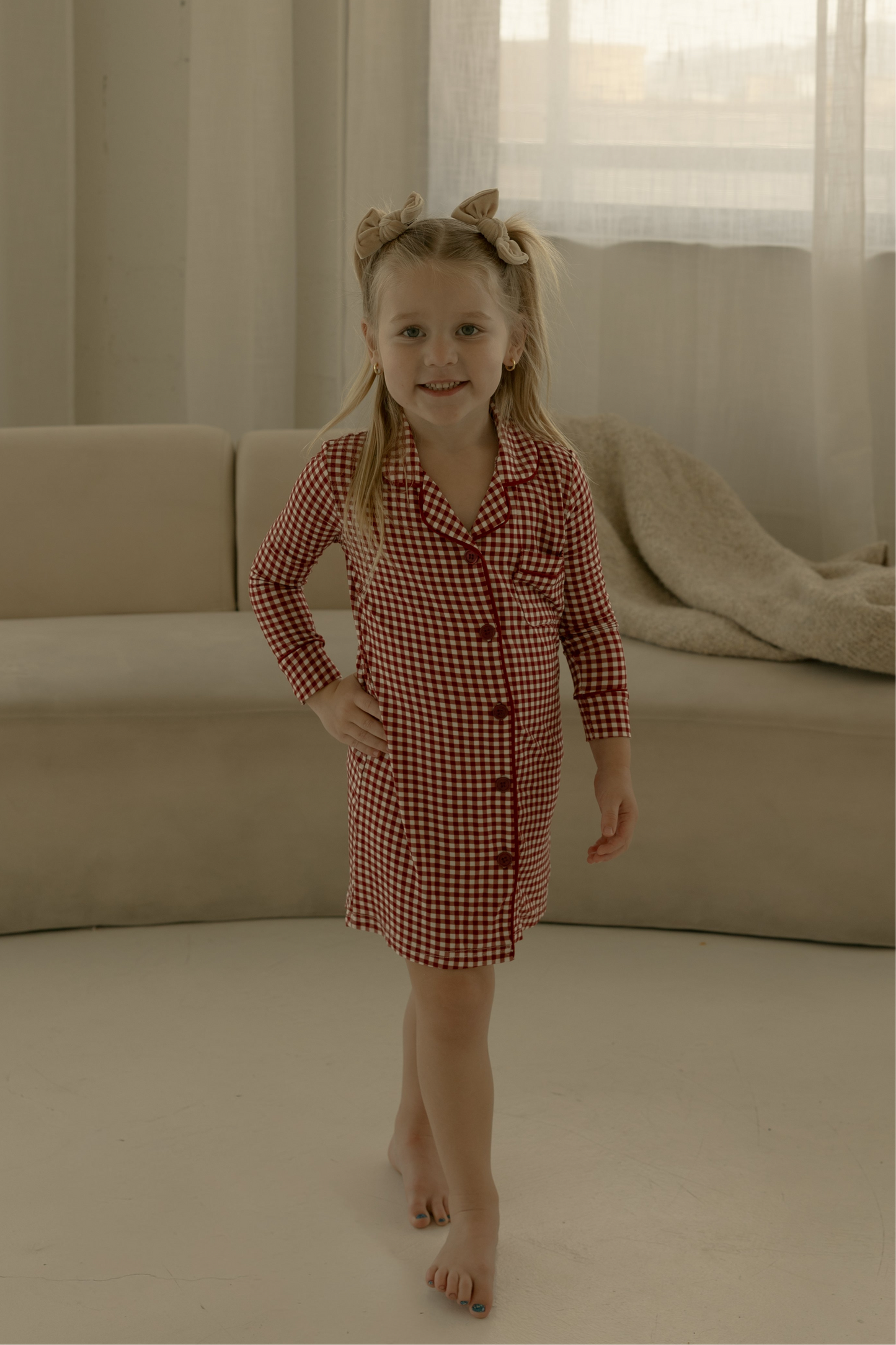 A young girl smiles barefoot indoors, wearing the forever french baby Toddler Bamboo Sleeping Dress in Red Gingham, standing before a beige sofa and large windows with sheer curtains.