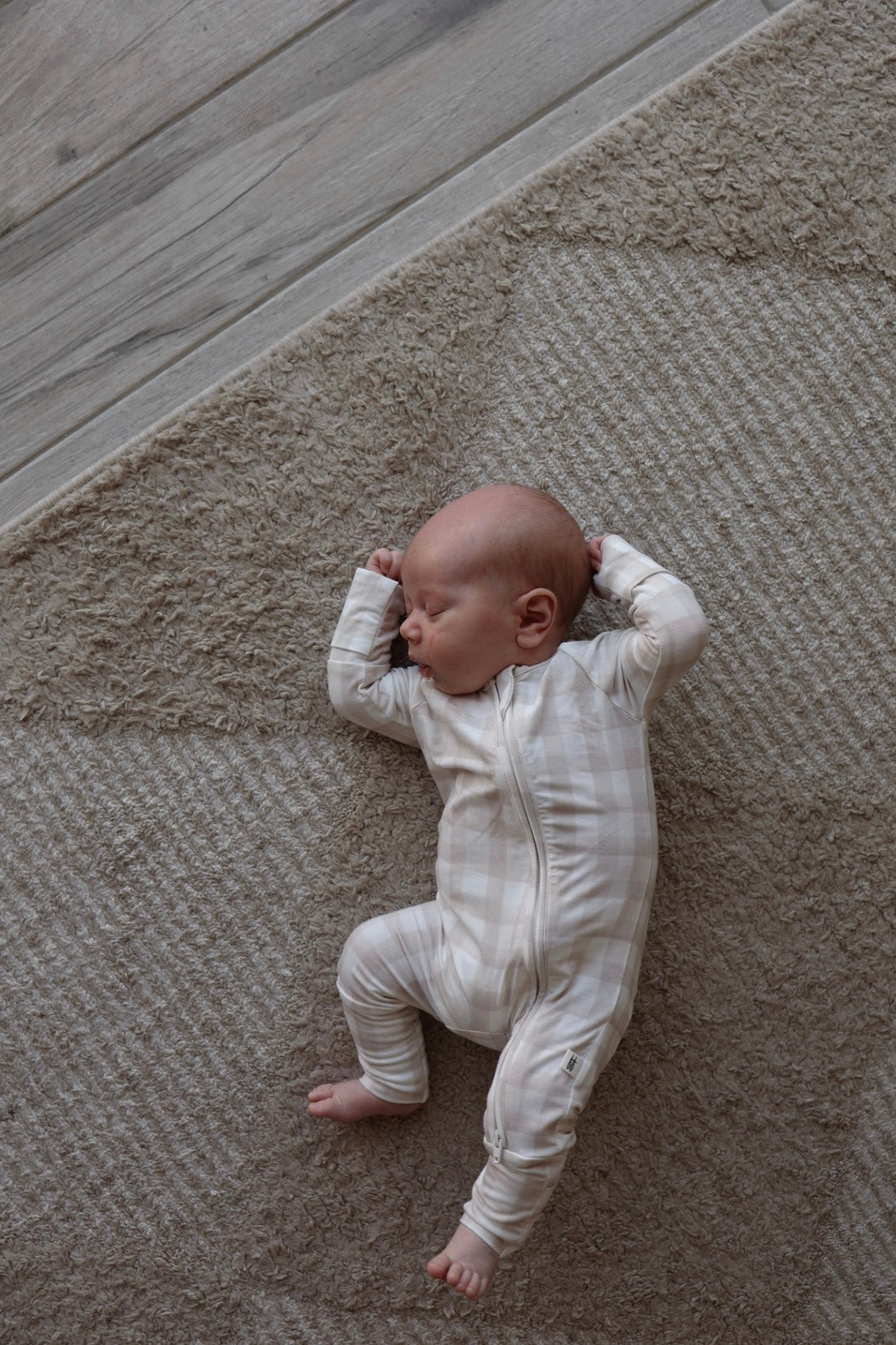 A baby in forever french baby Bamboo Zip Pajamas | Gingham, featuring a light plaid pattern, sleeps on a textured beige carpet beside a wooden floor, arms bent and hands near their head.