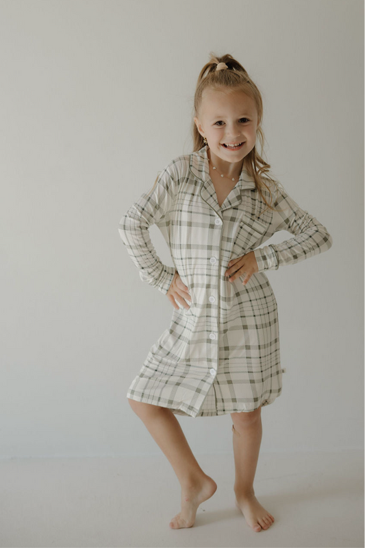 A smiling young girl with a high ponytail poses barefoot against a light background, wearing the forever french baby Toddler Bamboo Sleeping Dress in Picnic Plaid, her hands on her hips and one knee bent.