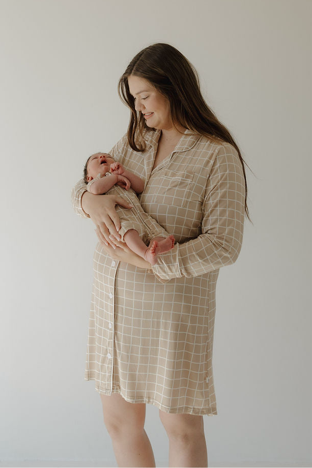 A smiling woman with long brown hair wears the forever french baby Women's Bamboo Sleeping Dress | Grid, holding a newborn in a light brown outfit against a plain, light background.