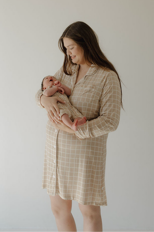 A smiling woman with long brown hair wears the forever french baby Women's Bamboo Sleeping Dress | Grid, holding a newborn in a light brown outfit against a plain, light background.