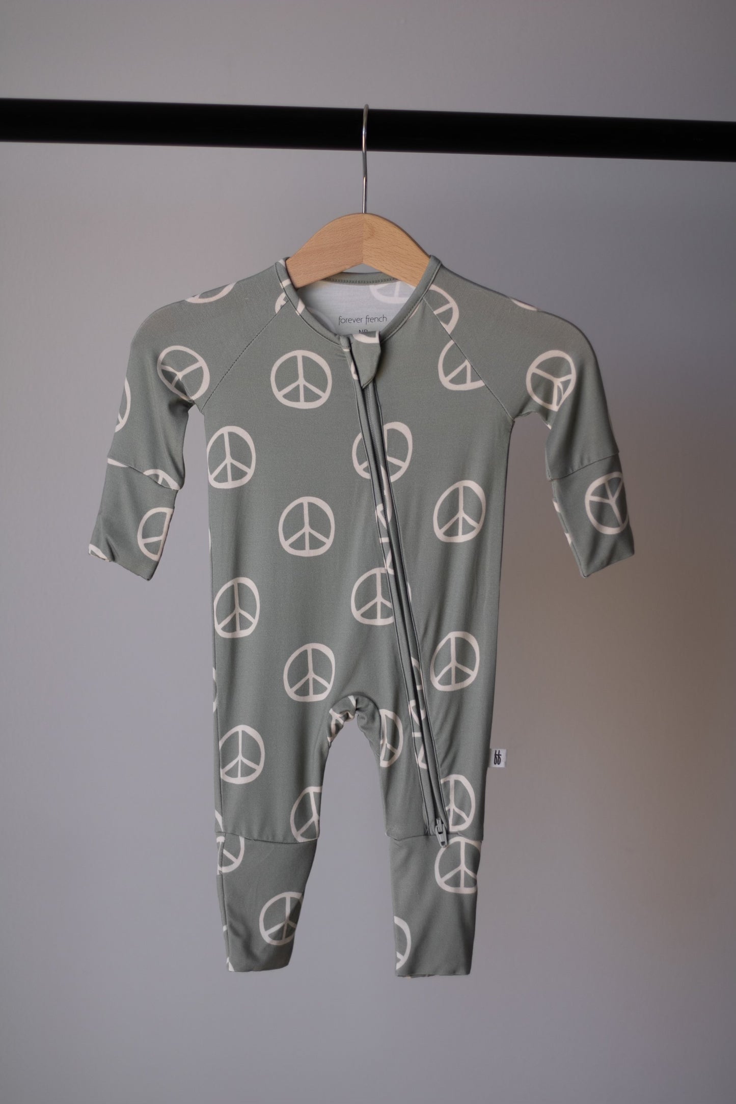 The Bamboo Zip Pajamas | Peace by forever french baby is a sage green, long-sleeve baby onesie featuring white peace signs, made from breathable fabric and shown hanging on a wooden hanger against a gray background.