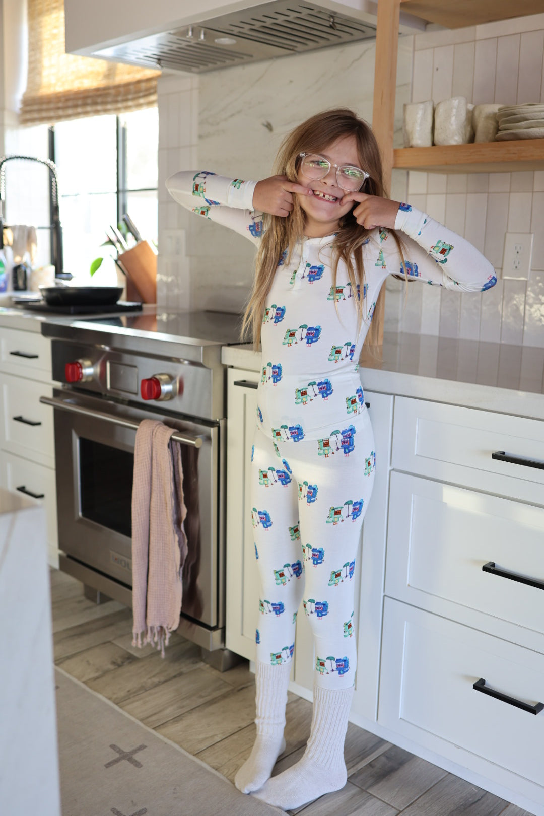 A young girl in forever french baby’s Bamboo Two Piece Pajamas | Charli's Monsters and glasses stands barefoot on a kitchen rug, smiling and pulling the corners of her mouth with her fingers near a stove and white cabinets.