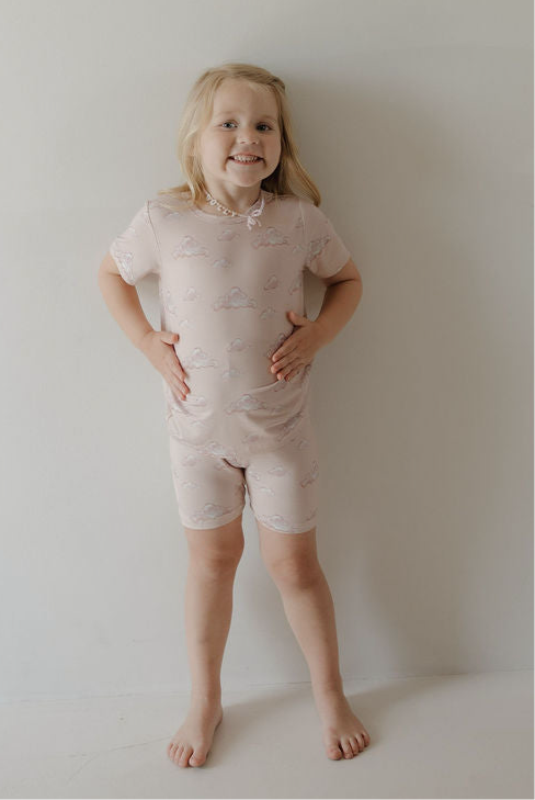 A smiling young child stands barefoot against a plain wall, wearing the Kendy 3 Bamboo Short Set | Ruby—crafted from breathable fabric for all-day comfort.