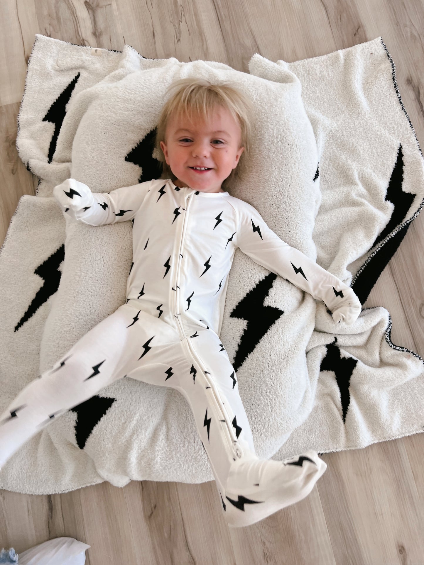 A smiling young child with light hair is lying on a cozy, white blanket adorned with black lightning bolt patterns. The child, wearing Forever French Baby's Bamboo Zip Pajamas in White & Black Lightning Bolt, stretches comfortably with arms and legs splayed.