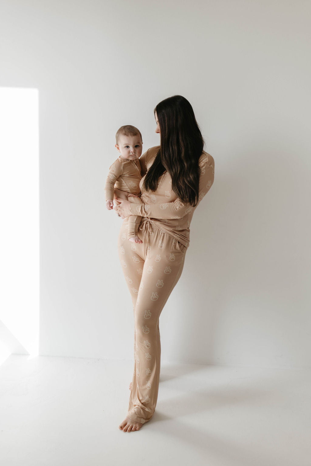 A woman in forever french baby Bamboo Zip Pajama | Peace Sign stands barefoot with her baby, who is also in matching breathable pajamas, both gazing calmly at the camera against a plain white background.