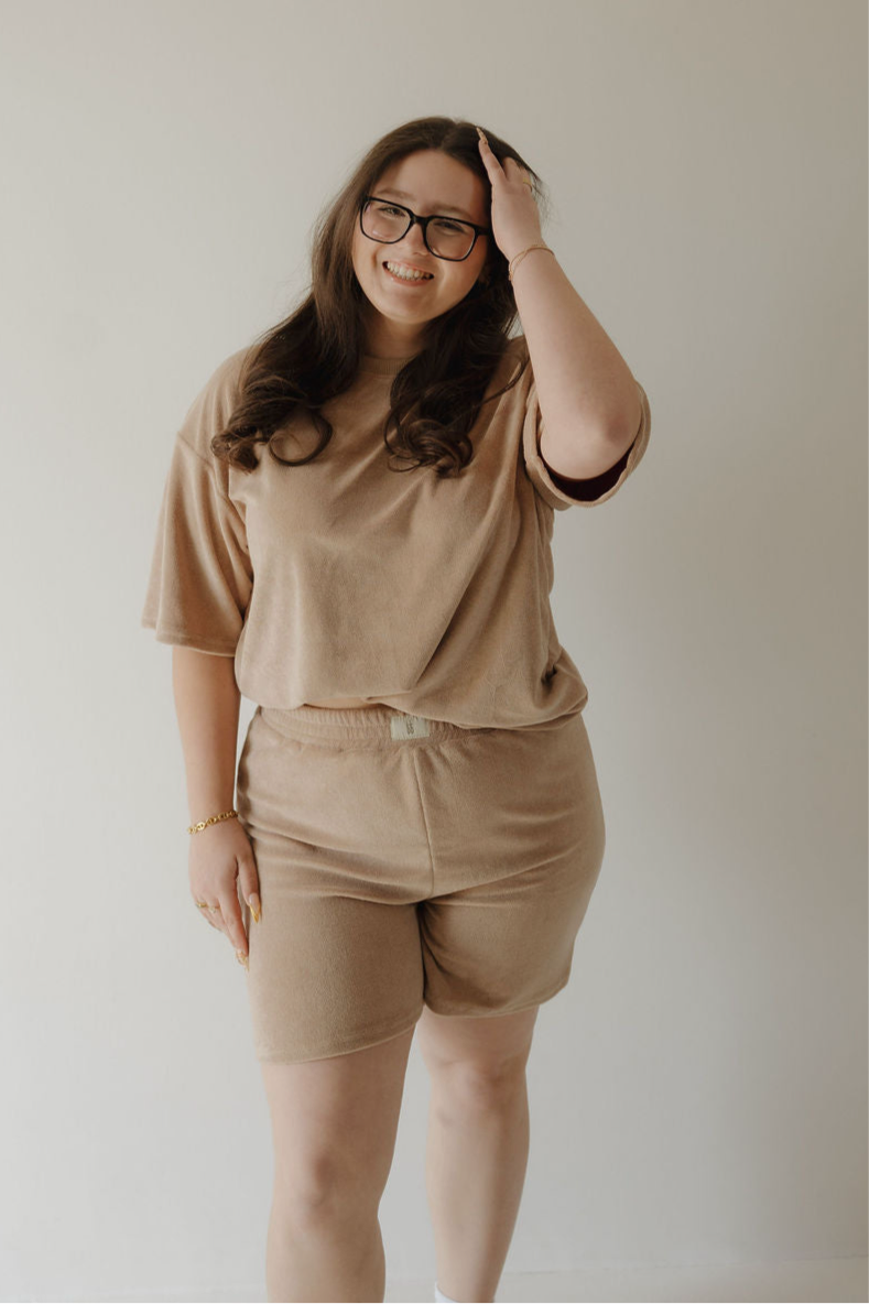 A woman with long brown hair and glasses smiles while touching her head. She wears the forever french baby Women's Terry Short Set in Tan, standing against a plain light-colored background.