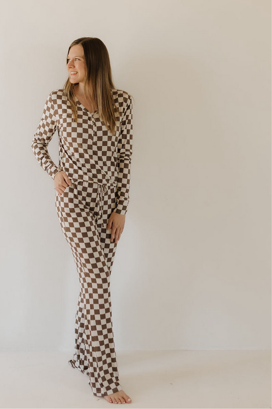 A woman smiles against a plain wall, wearing the forever french baby Women's Bamboo Pajama | Dutton—a brown and white checkerboard long-sleeve set with flared pants, crafted from breathable fabric.