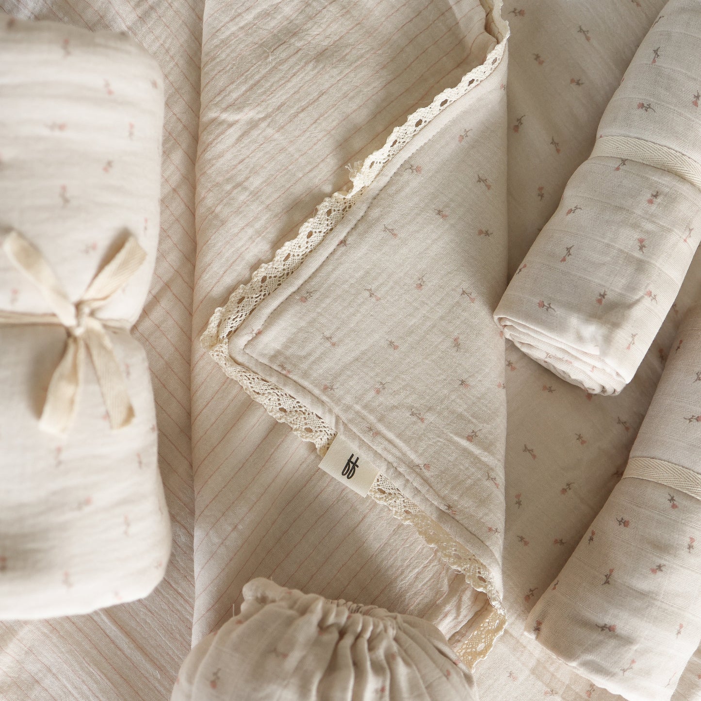 The forever french baby Muslin Swaddle | Lucy, in soft cream with delicate lace trim and subtle floral patterns, is neatly rolled and arranged on a matching fabric background. One swaddle displays a small fabric tag.