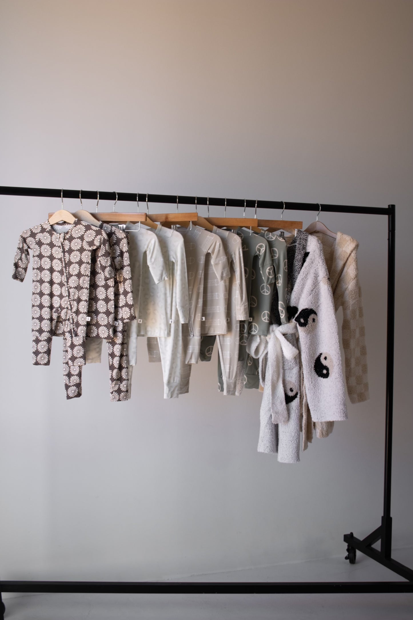 A clothing rack displays seven sets of forever french baby Bamboo Two Piece Pajamas | Peace and a robe, in neutral and pastel patterns like flowers, checks, and yin-yangs, all hanging neatly on wooden hangers against a plain wall.