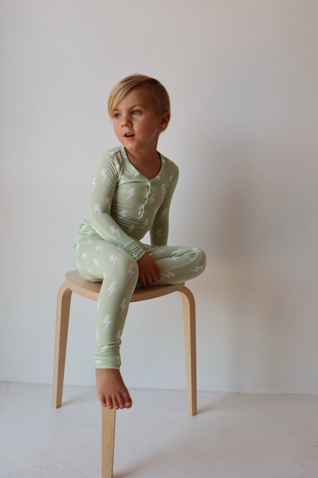 A young child with short blond hair sits cross-legged on a light wooden stool, wearing forever french baby's Bamboo Two Piece Pajamas | Flash in light green with a white lightning bolt pattern—perfect breathable sleepwear for all-night comfort.