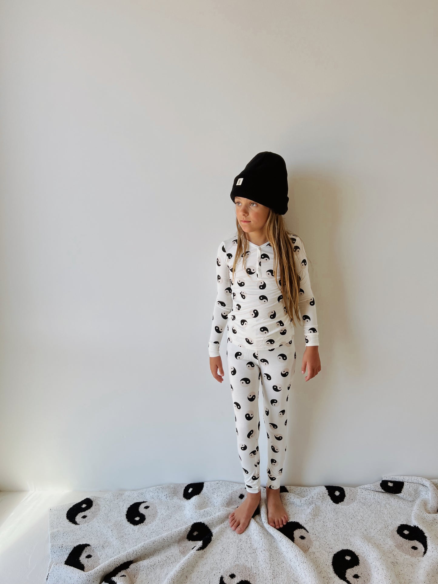 A young girl stands barefoot on a white rug patterned with yin-yang symbols, wearing a Bamboo Two Piece Pajamas | Black & White Yin Yang from forever French baby. Her long hair is loose under a black beanie hat as she gazes to the side in front of a plain white wall. The hypo-allergenic clothing ensures she feels comfortable as well as stylish.