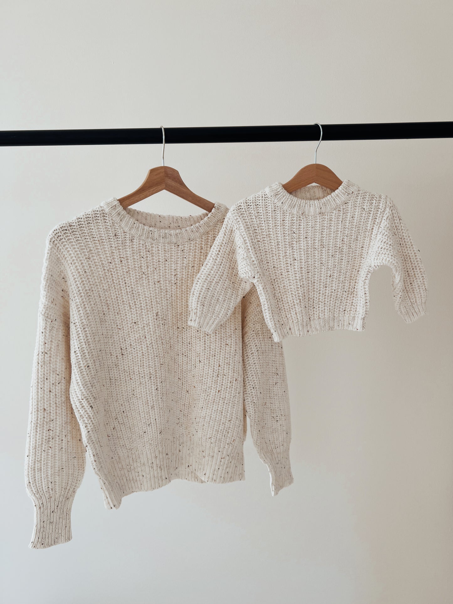 Two cream child-sized and adult-sized knit sweaters by forever french baby, in Cloud Confetti, hang on wooden hangers against a plain wall, creating a cozy, coordinated look.