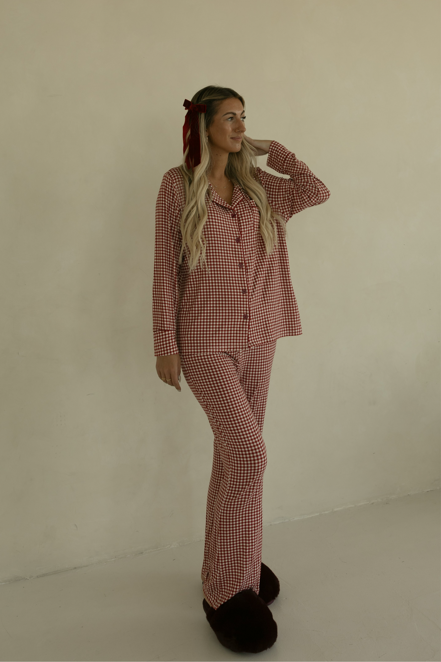 A woman with long blonde hair smiles to the side while wearing forever french baby's Long Sleeve Women's Bamboo Pajamas in Red Gingham, a soft, hypo-allergenic lounge set, paired with dark slippers against a plain beige wall.