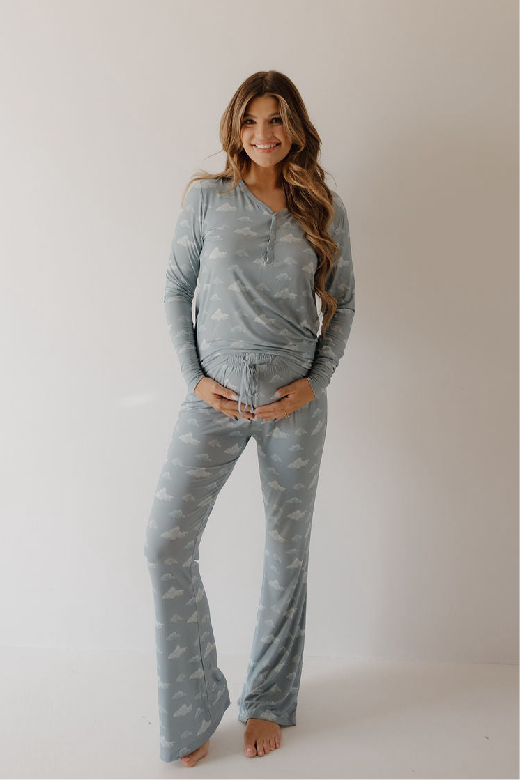 A smiling woman with long hair stands barefoot against a plain background, wearing Kendy 3’s Women's Bamboo Pajama | Duke in a light blue cloud pattern. She rests her hands on her belly, enjoying the breathable bamboo fabric.