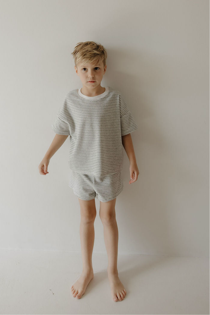 A young boy with short blonde hair stands barefoot against a plain, light wall, wearing the forever french baby Child Terry Short Set in Grey Stripe—perfect for cozy family moments.