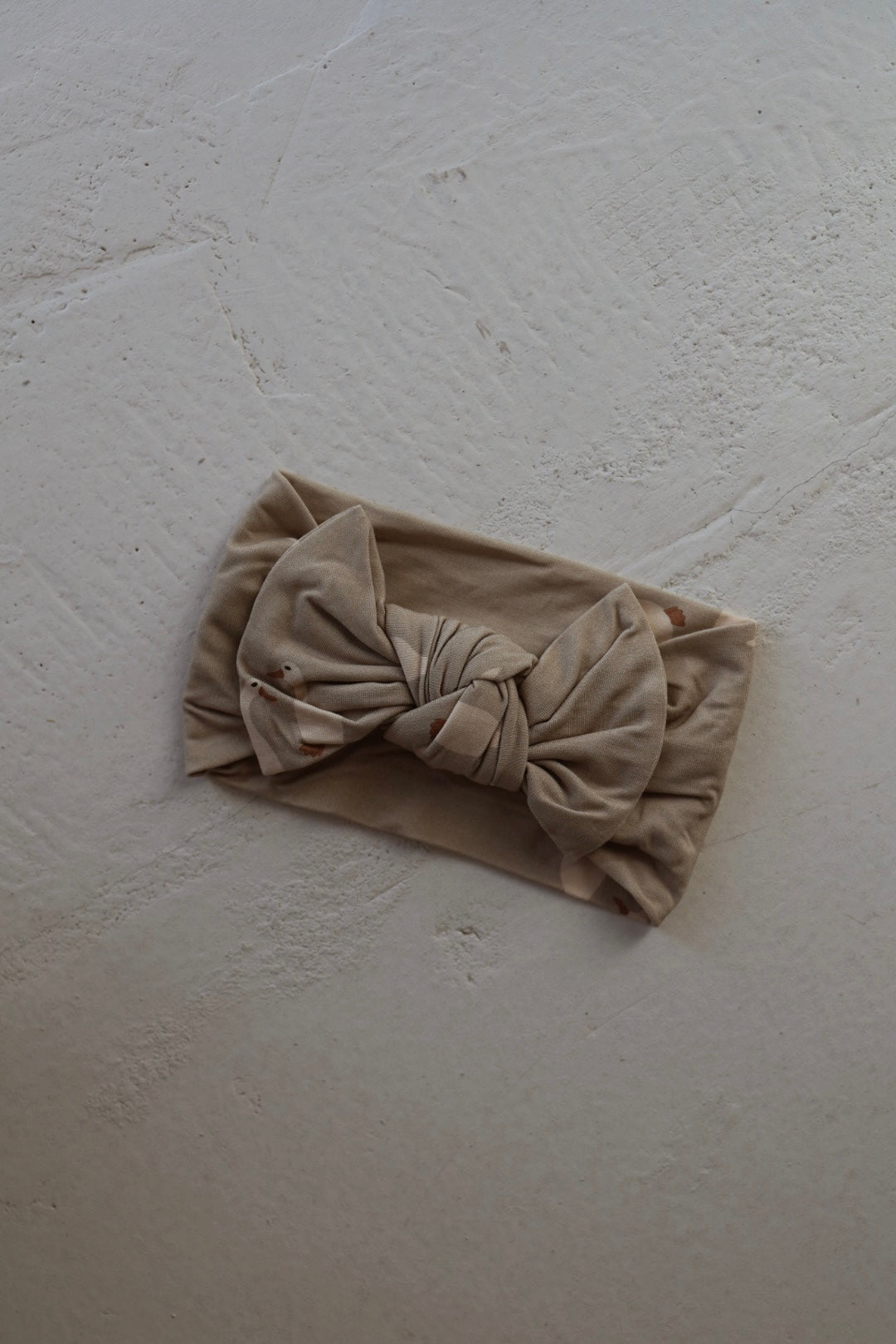 The Bamboo Head Wrap from forever french baby by Silly Goose, in beige with a large bow, rests on a textured light surface.