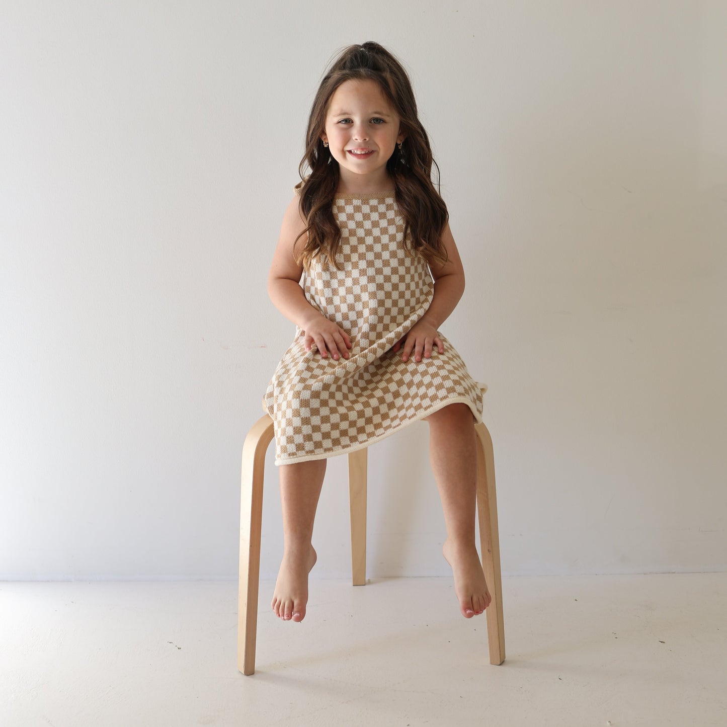 A young girl with long brown hair, barefoot and smiling, sits on a wooden chair against a plain white wall, wearing the Knit Dress | Latte Check from forever french baby.