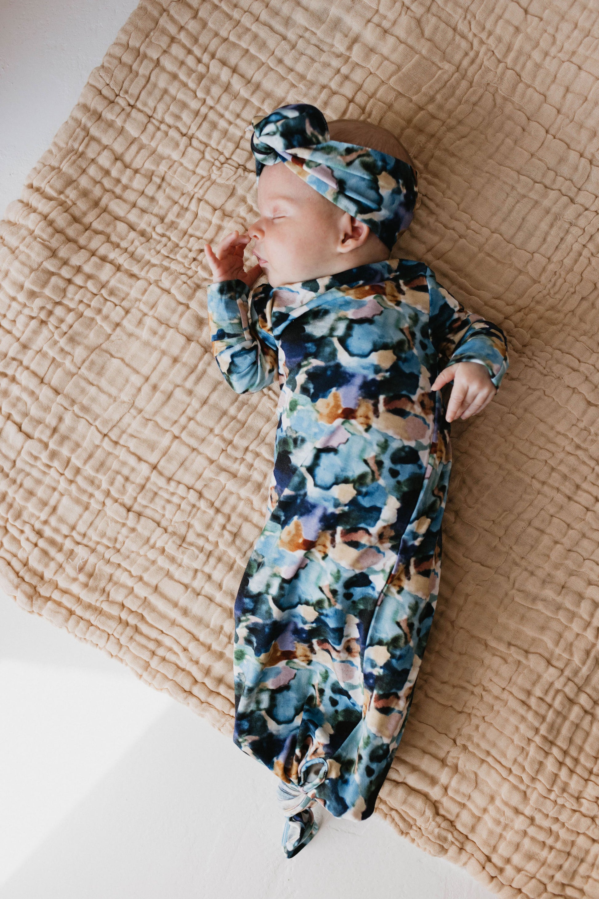 A baby sleeps on a beige, textured blanket, wearing a colorful floral-patterned Bamboo Knotted Gown in the Charli Print by Forever French Baby. With a matching headband and their left hand near their face, the baby appears to be peacefully resting—a serene moment that could inspire autistic daughter art.