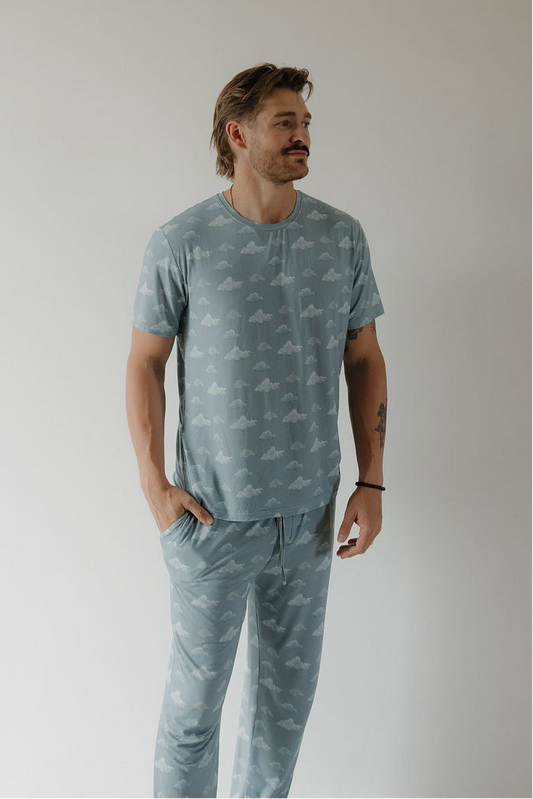 A man with light brown hair and a mustache stands against a white background, wearing the Kendy 3 Men's Short Sleeve Bamboo Pajama | Duke, featuring a cloud pattern. With his left hand in his pocket, he smiles in the breathable fabric.