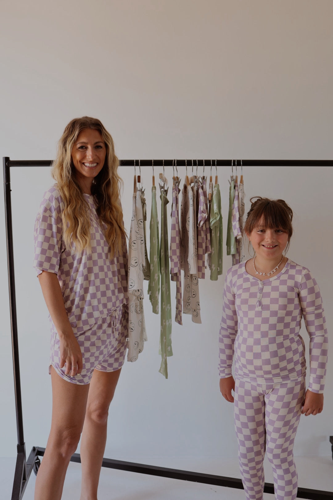 A woman and a girl, both in matching forever french baby's Women's Bamboo Short Set in Grape Soda, stand by a clothing rack with patterned garments against a plain background.
