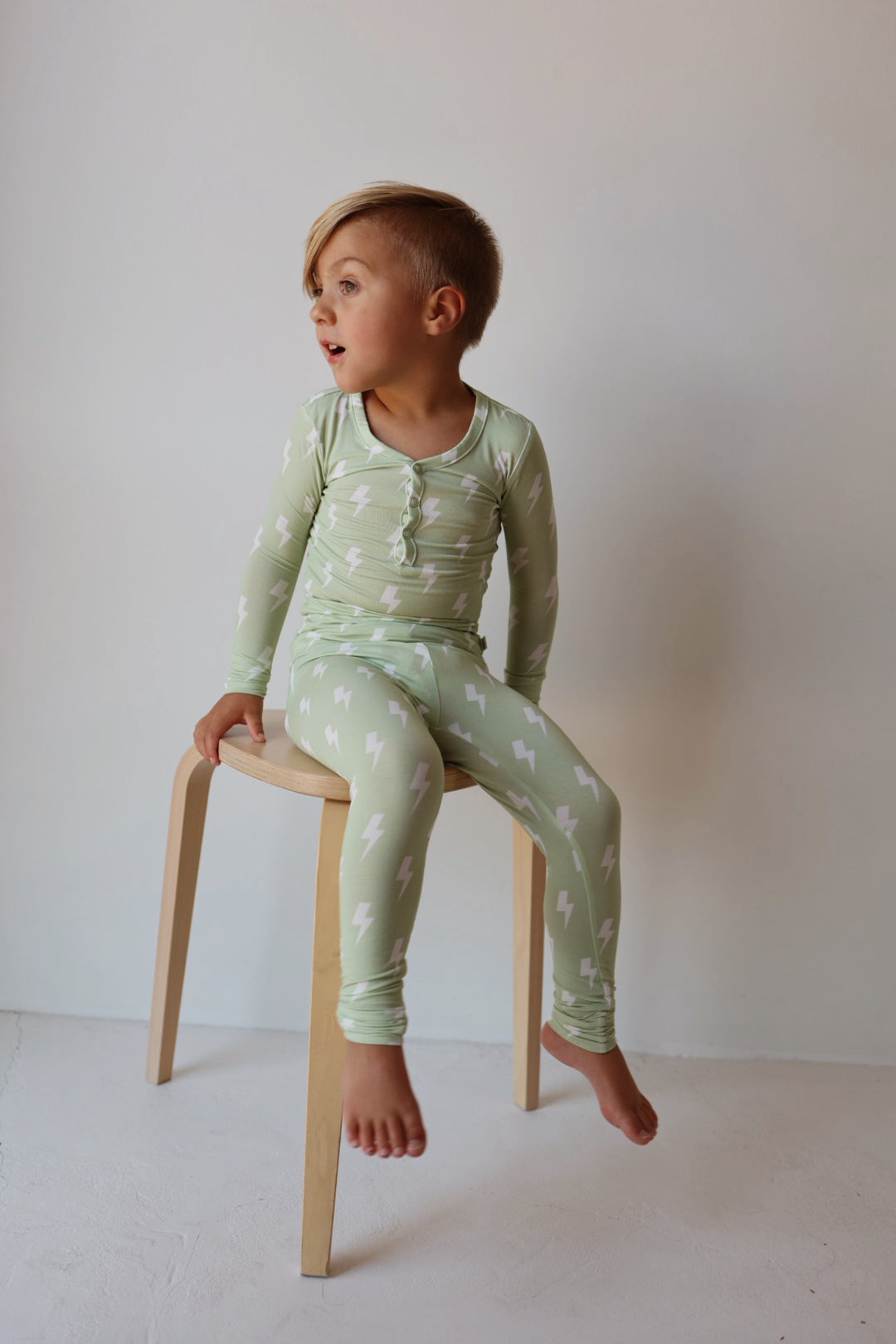 A young child with short blonde hair sits barefoot on a wooden stool, wearing forever french baby’s Bamboo Two Piece Pajamas | Flash—light green sleepwear with a white lightning bolt pattern—against a plain light background.