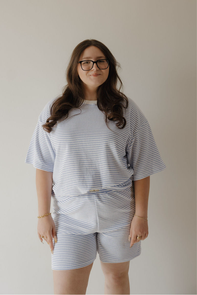 A woman with long brown hair and glasses stands against a plain background, wearing the forever french baby Women's Terry Short Set in Ocean Stripe—ideal for effortless, everyday style. She smiles slightly with her hands relaxed at her sides.