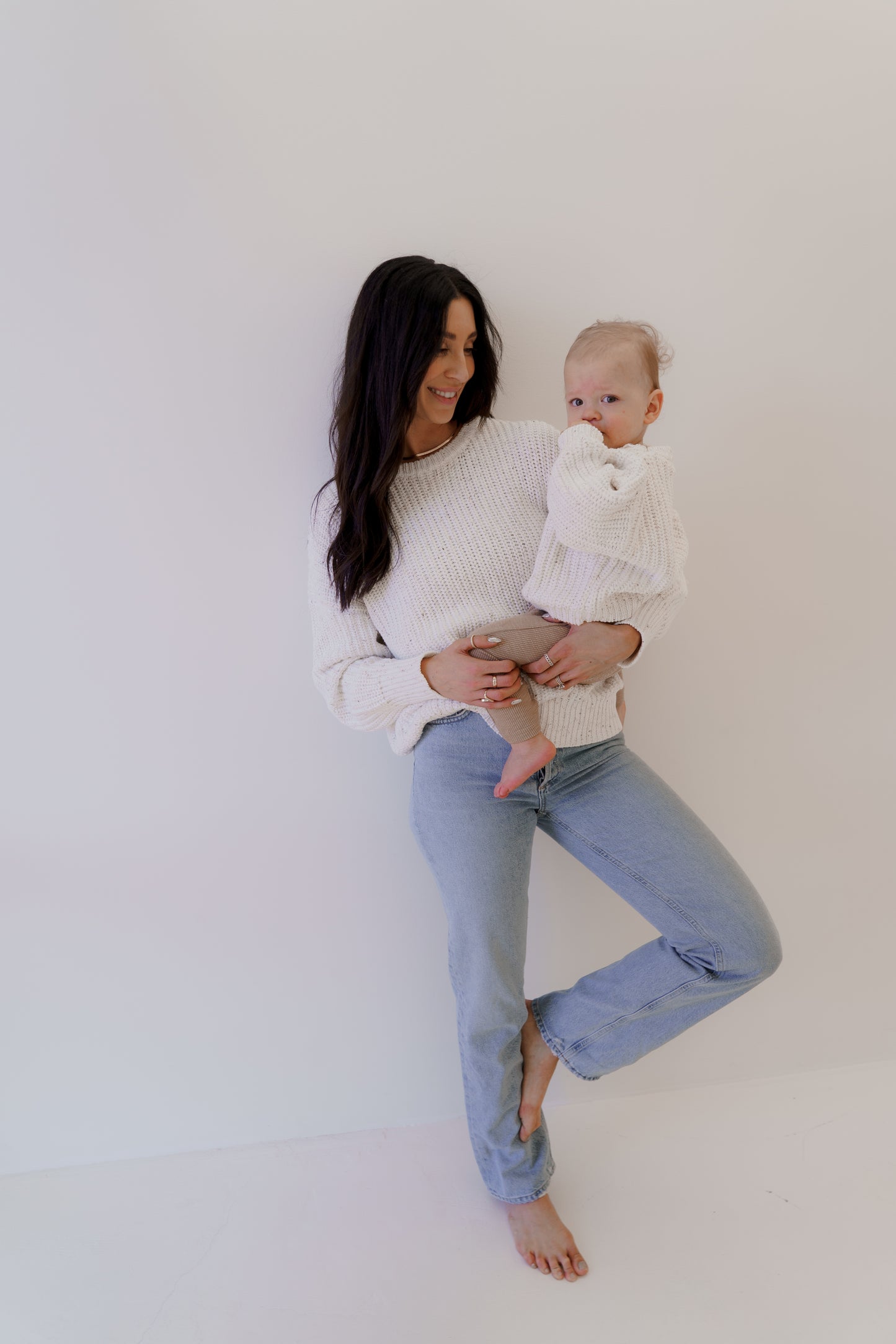 A woman with long dark hair stands against a white wall, holding a baby. They're both wearing white sweaters and jeans. The woman is smiling while the baby, wearing a "Child Knit Sweater | Cloud Confetti" by forever french baby, looks at the camera, sucking their thumb.
