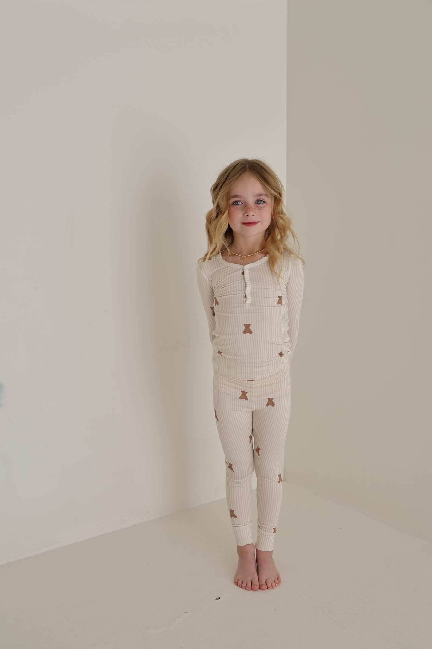 A young girl with blonde wavy hair smiles gently, standing against a white wall with her hands behind her back. She wears forever french baby’s Bamboo Two Piece Pajamas | Teddy—cream, hypoallergenic sleepwear adorned with a brown teddy bear print.