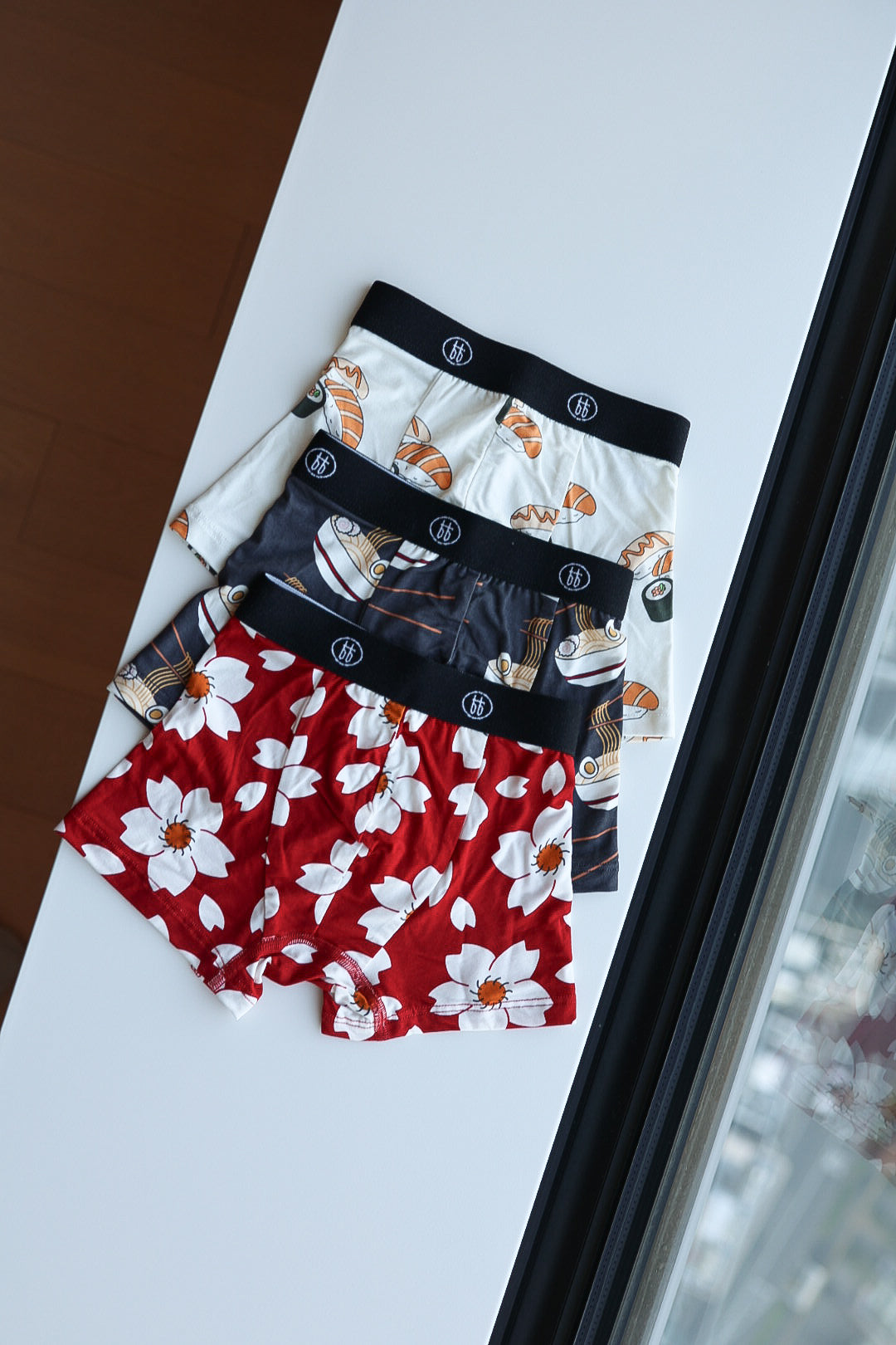 Three pairs of forever french baby Bamboo Boxers (3 Pack)—featuring Cherry Blossoms, Ramen, and Sushi patterns—are neatly folded on a white surface by a window.