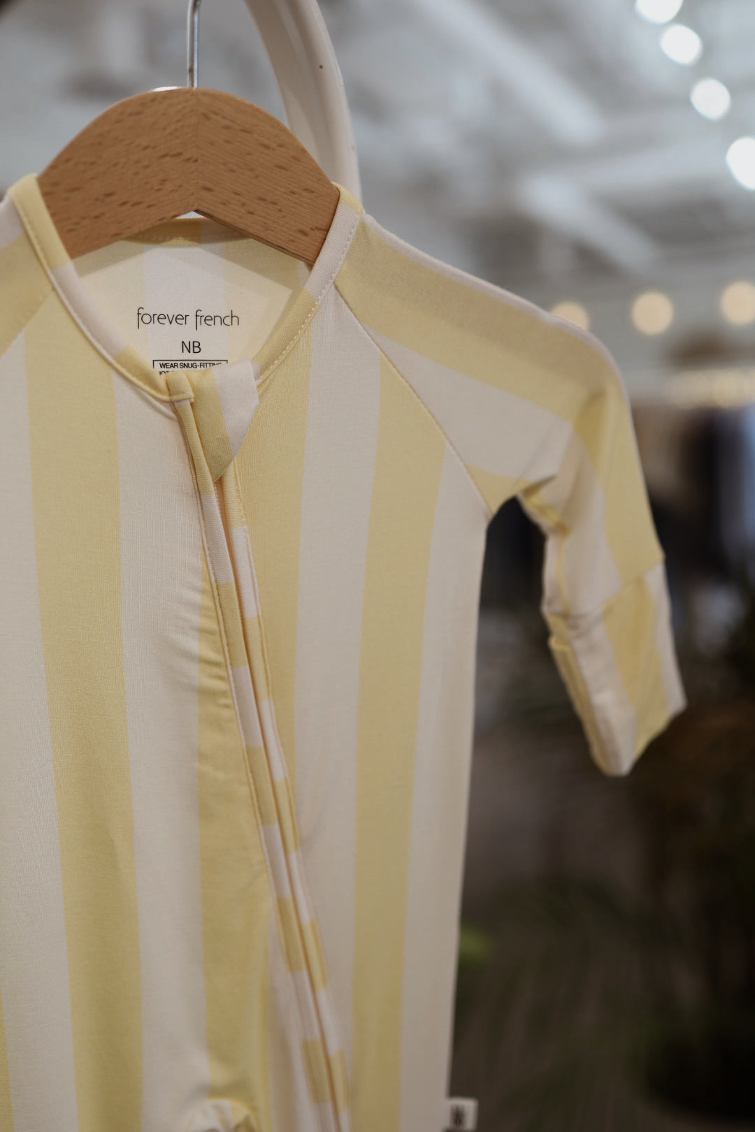A yellow and white striped Bamboo Zip Pajamas | Butter by forever french baby hangs on a wooden hanger with an NB size tag, photographed indoors against a softly blurred background.