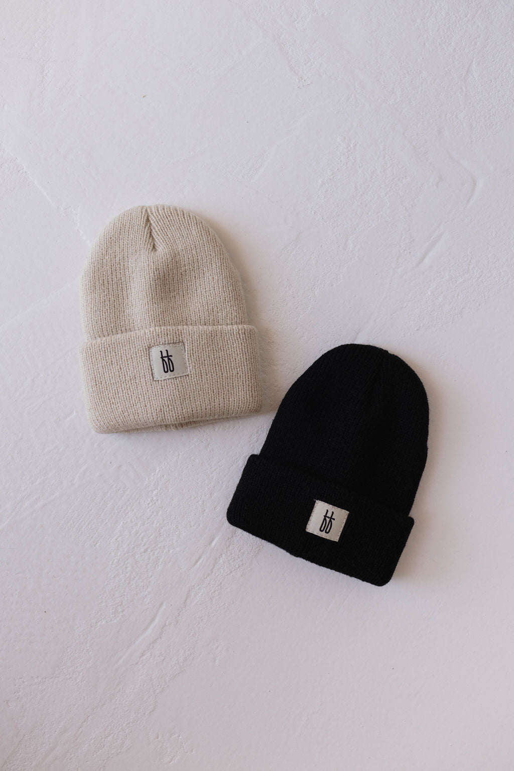 A beige and a black ff Knit Beanie by forever french baby, both featuring ribbed designs, rest on a white textured surface. Each beanie displays a small, rectangular white patch adorned with a minimalist black monogram logo.