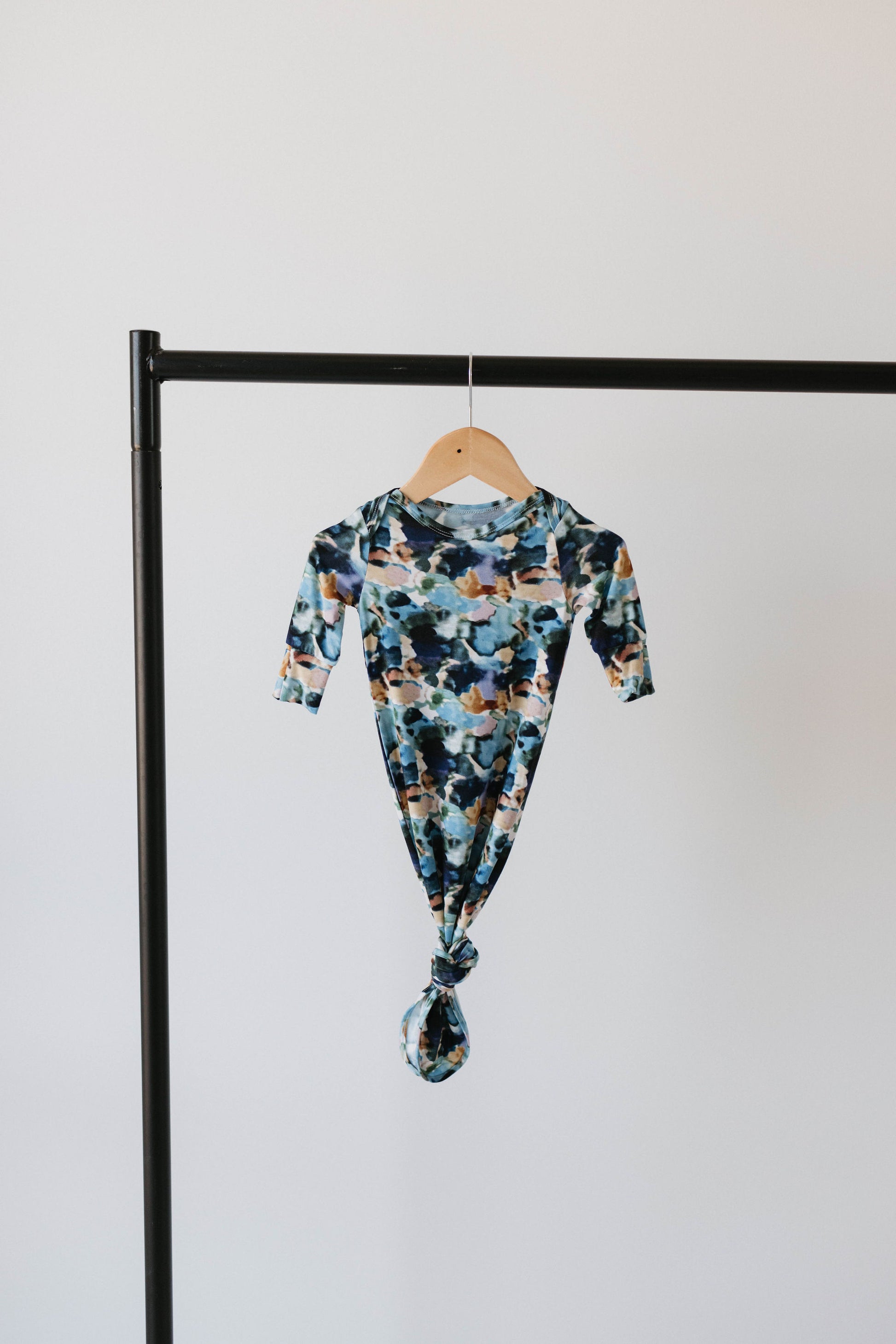 A "forever french baby" Bamboo Knotted Gown in the Charli Print, featuring a colorful abstract pattern primarily in blue, black, and beige hues, hangs on a wooden hanger from a black metal clothing rack. The bottom of the gown is tied in a knot. The background is a plain white wall, reminiscent of the soothing hues often found in autism awareness themes.