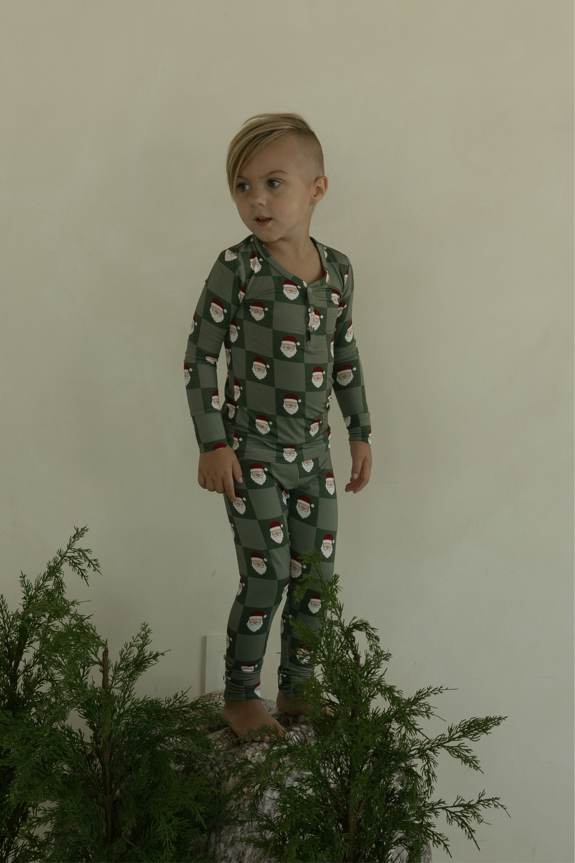 A young child with short, side-swept blonde hair stands on a rock in forever french baby’s Bamboo Two Piece Pajamas | Santa Checkerboard, surrounded by green foliage and set against a plain light-colored wall.