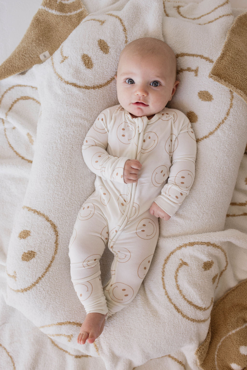 An infant, dressed in Forever French Baby's Bamboo Zip Pajamas | Just Smile, featuring cheerful smiley face prints, lies on a blanket adorned with the same design. The baby gazes up with wide eyes, one hand resting on their stomach. The soft and cozy background creates a warm atmosphere, perfect for hypoallergenic clothing.