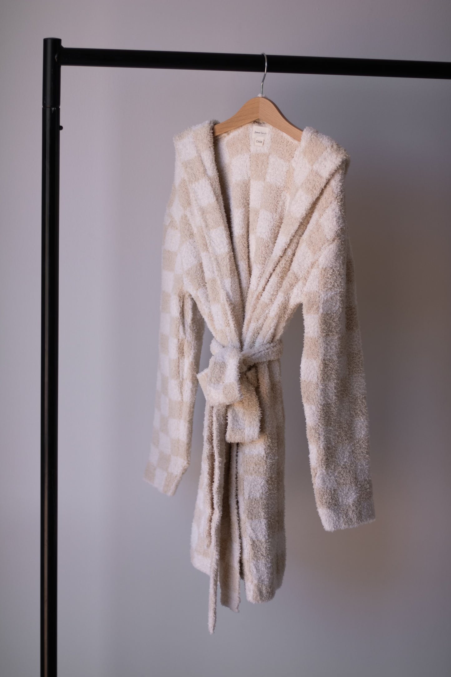 A forever french baby Adult Robe in Cream Checkerboard hangs on a wooden hanger, displayed on a black metal clothing rack against a plain light wall.