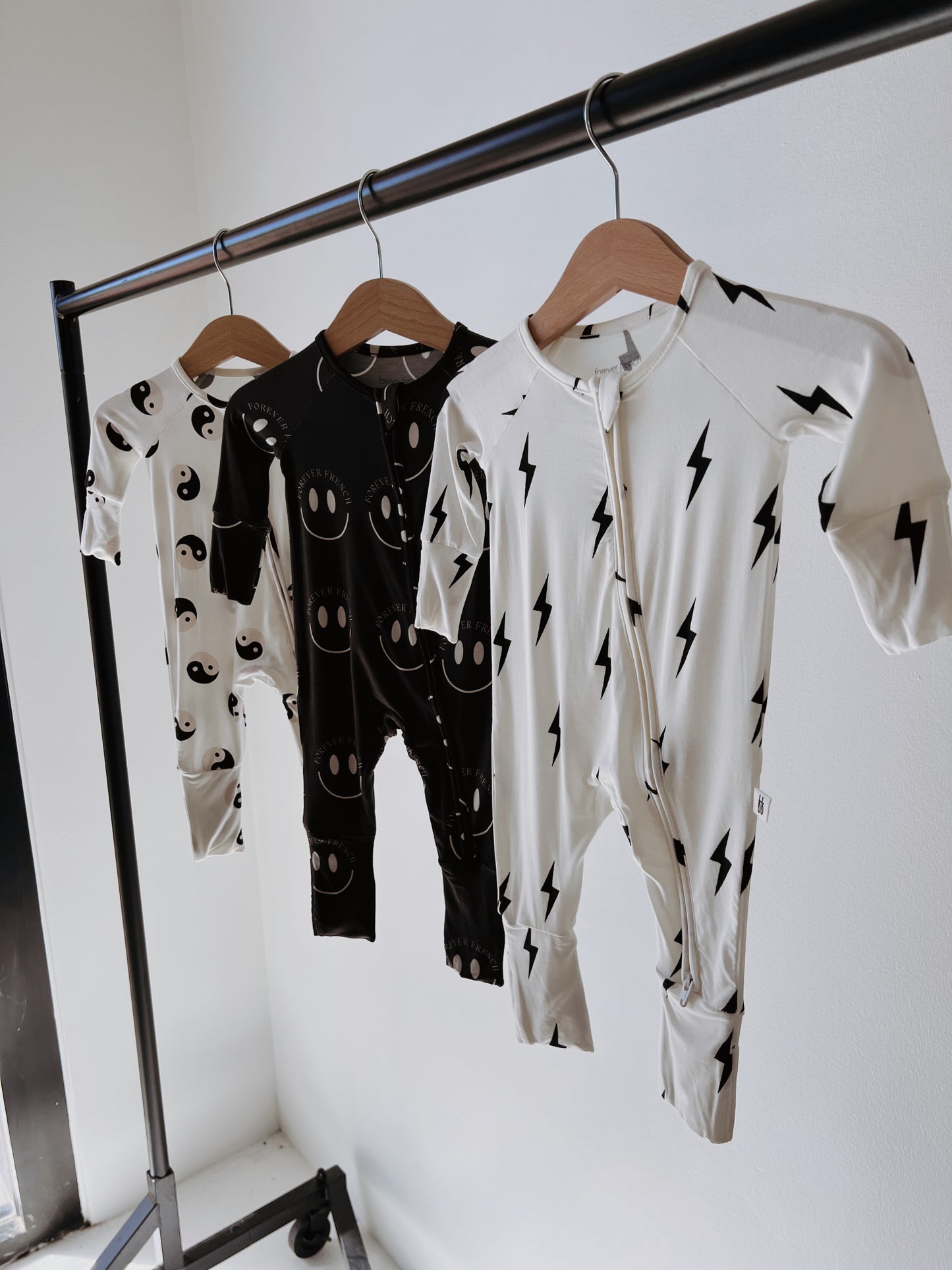 Three baby and toddler Bamboo Zip Pajamas from forever french baby are displayed on a metal clothing rack. The pajama on the left features a black fabric with white smiley faces, the middle one showcases black with white lightning bolts, and the right one renders a striking yin yang design in white with black lightning bolts.