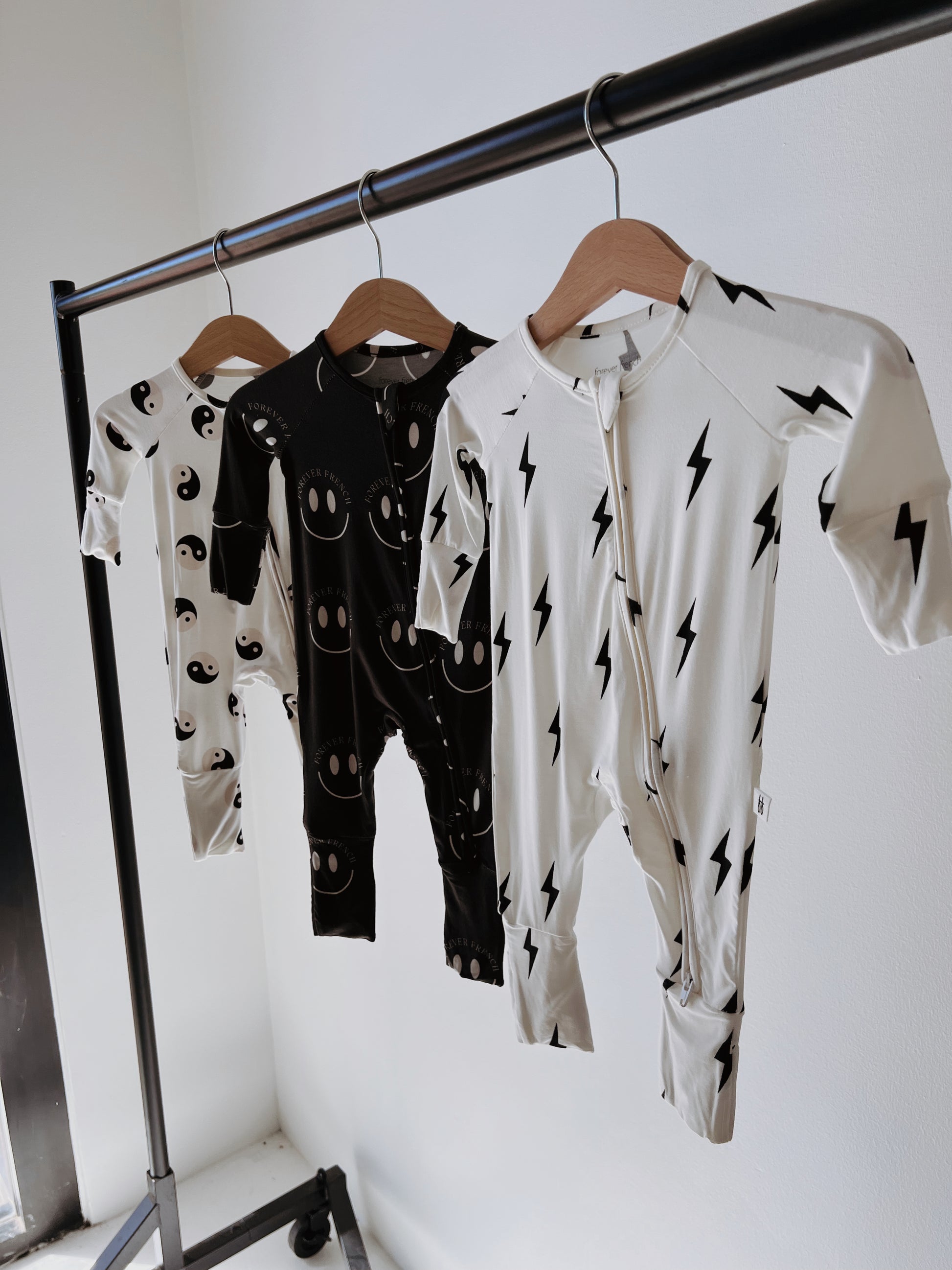 Three baby and toddler Bamboo Zip Pajamas from forever french baby are displayed on a metal clothing rack. The pajama on the left features a black fabric with white smiley faces, the middle one showcases black with white lightning bolts, and the right one renders a striking yin yang design in white with black lightning bolts.