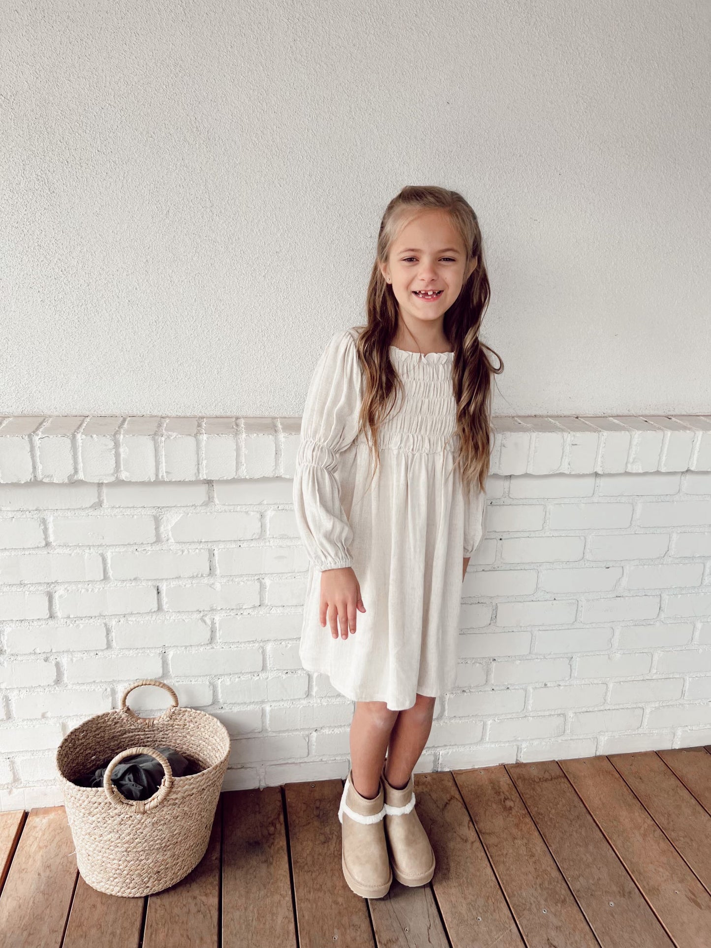A young girl with long brown hair stands smiling against a white brick wall. She is wearing the Women's Linen Dress | Flax from forever french baby and beige boots. A wicker basket is on the wooden floor beside her.