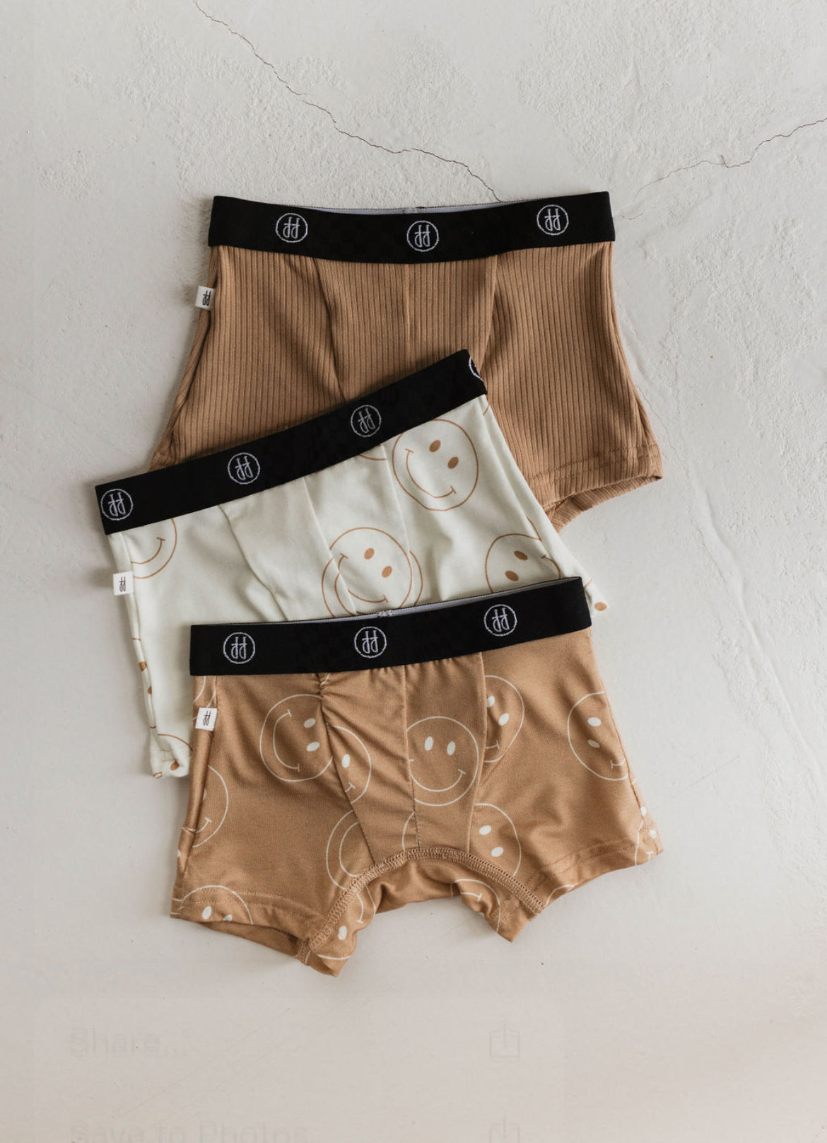 Three pairs of Bamboo Boxers (3 Pack) by forever french baby are arranged in a staggered stack on a white surface. The top pair is solid brown, the middle pair is off-white with smiley face prints, and the bottom pair is light brown with large smiley face prints. Made from hypo-allergenic bamboo fabric, these undies feature black elastic waistbands.