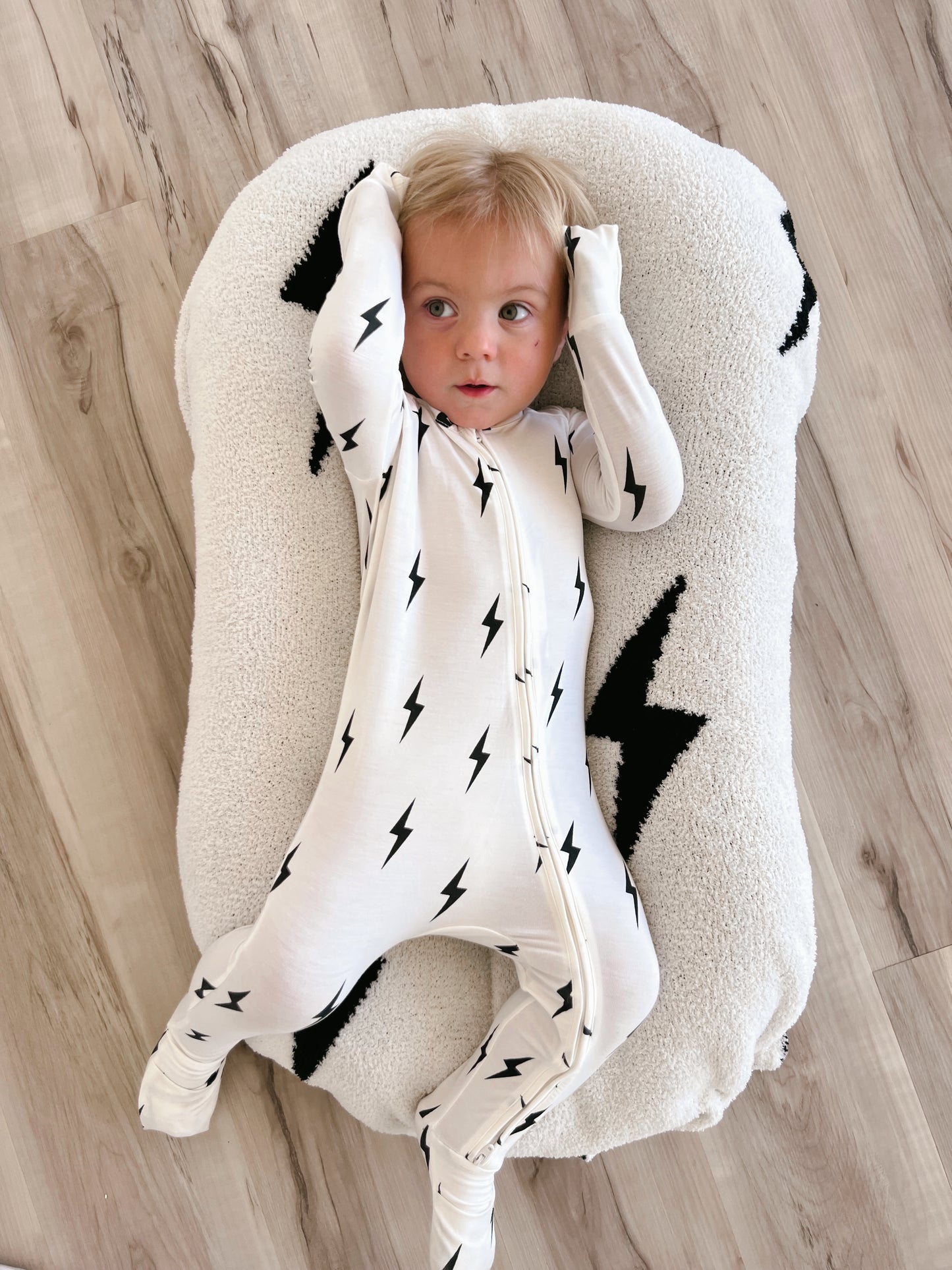 A baby wearing the Bamboo Zip Pajamas in White & Black Lightning Bolt by forever french baby lays on a plush, white pillow with matching lightning bolt designs. The baby's hands are up near their head as they look off to the side, with the soft background highlighting light-colored wooden flooring.