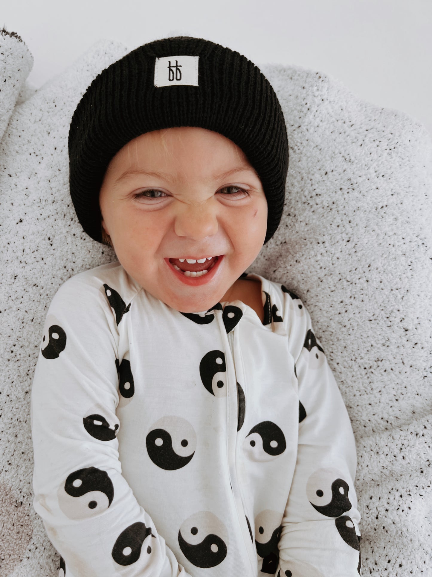 A baby wearing a black beanie with a logo and the forever french baby Bamboo Zip Pajamas in Black & White Yin Yang design is sitting on a soft gray blanket. This adorable piece from our Baby and Toddler clothing line features the Yin Yang design, perfect for your little one's stylish comfort.