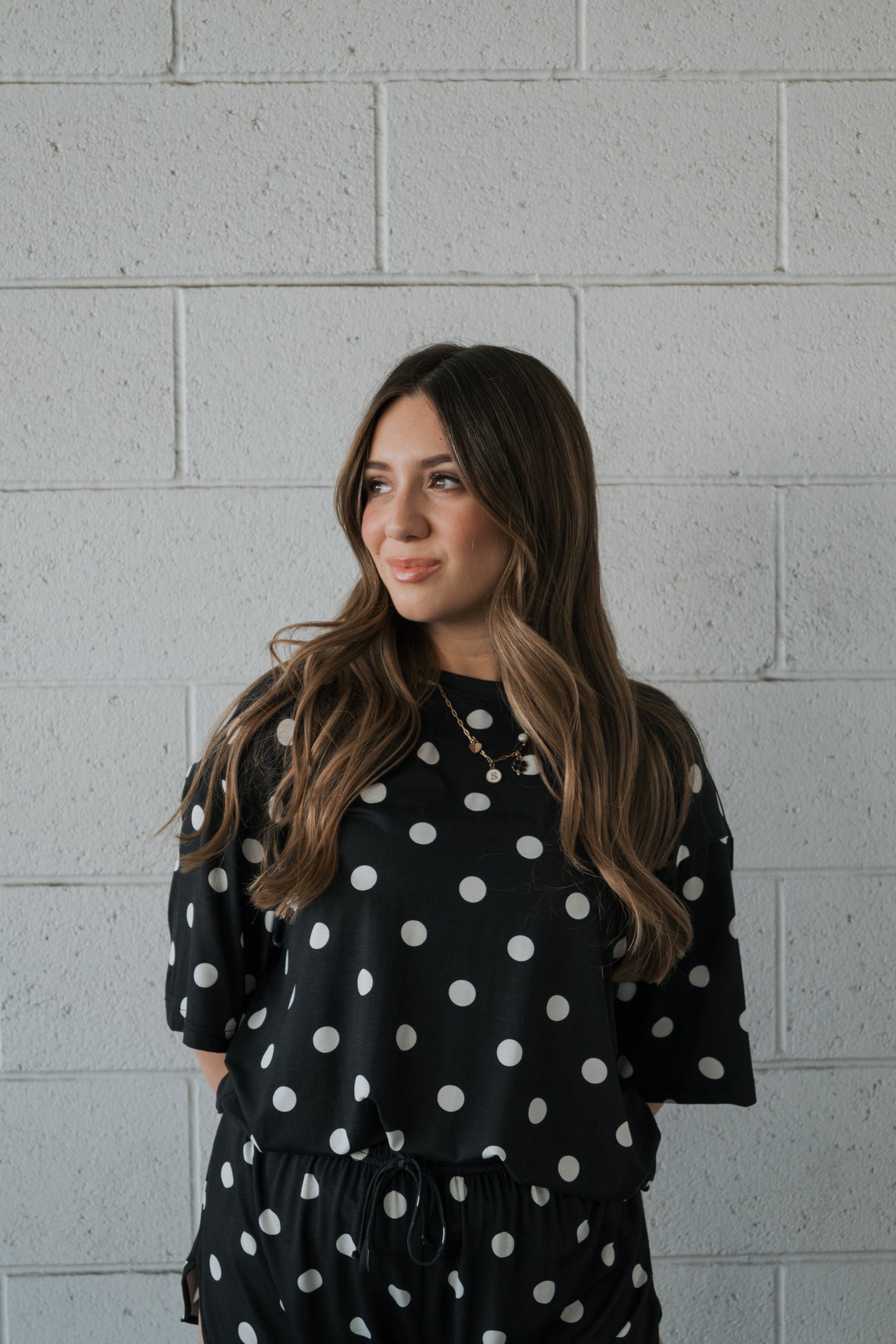 A woman with long brown hair stands against a light gray brick wall, smiling slightly to her left as she models the forever french baby Women’s Bamboo Short Set in the B&W Polka Dot print.