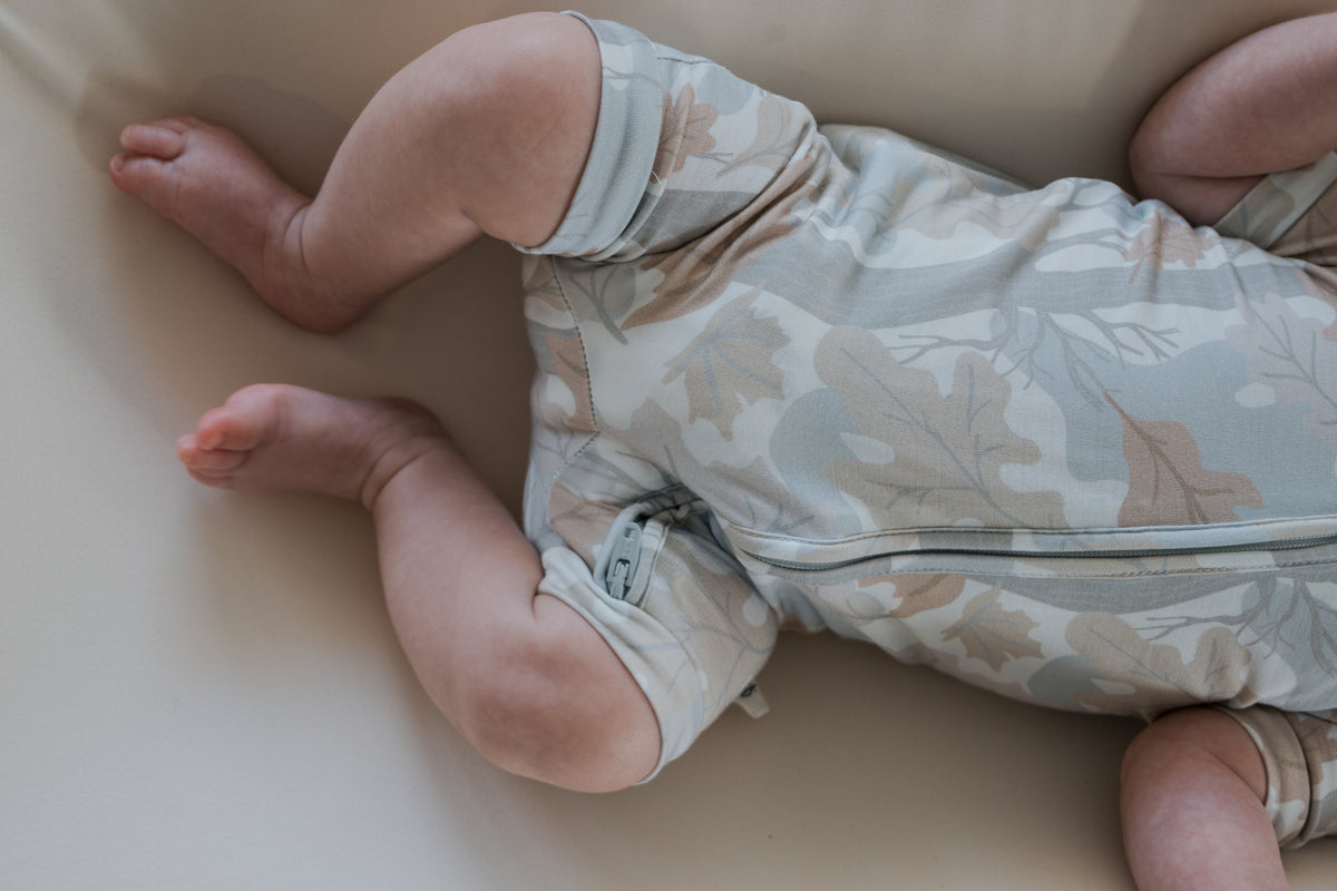 A baby wearing the forever french baby “Shortie Bamboo Zip | Woodland” romper, featuring a light woodland leaf pattern, lies on a soft, cream surface. Only the legs and part of the torso are visible, drawing attention to the gentle design and cozy comfort of this hypoallergenic sleepwear.