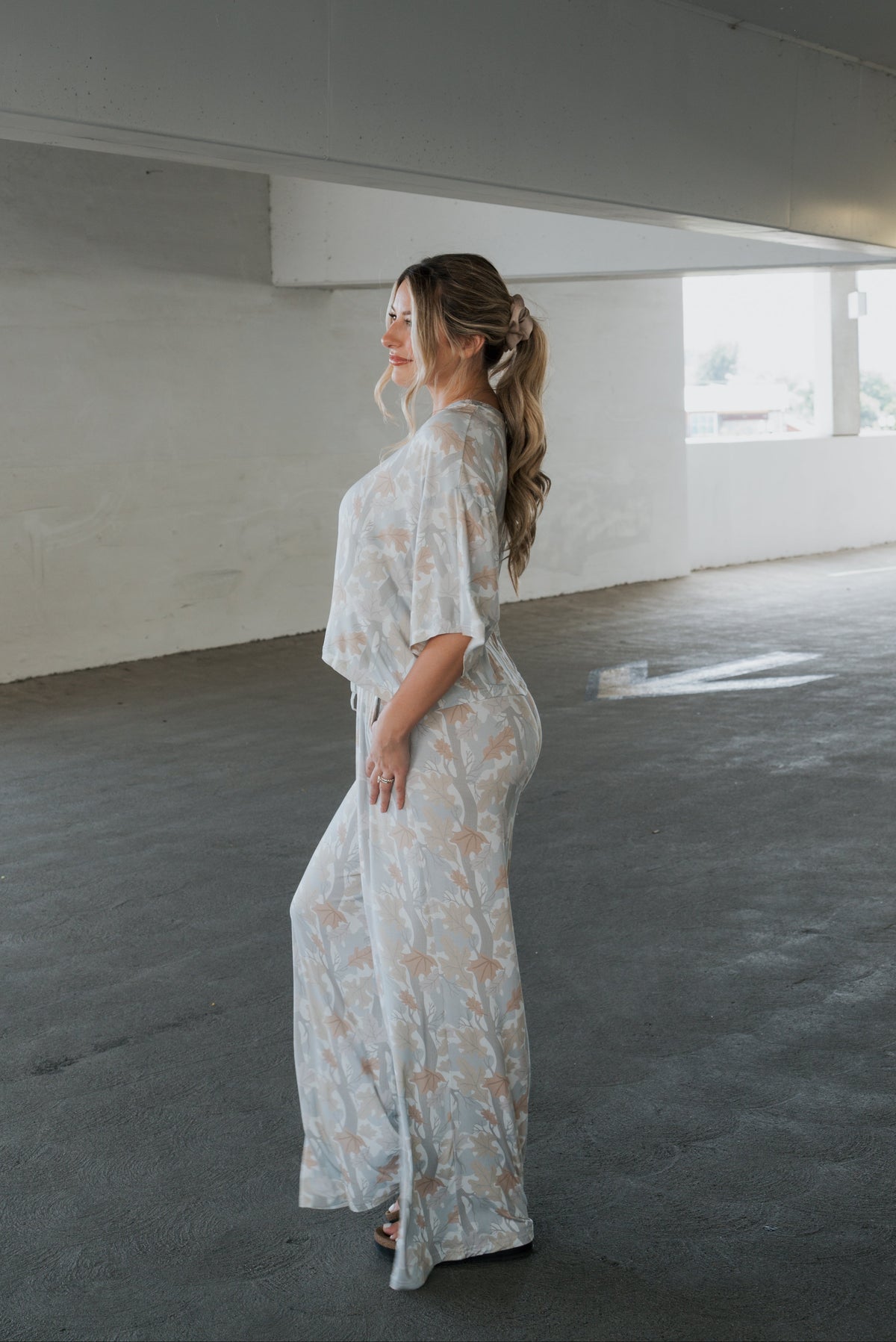 A woman stands in a parking garage wearing the forever french baby Women’s Bamboo Crop Tee Lounge Set in Woodland, a loose, light-colored floral outfit with wide-leg pants. Her long, wavy hair is tied back as she looks to the side, smiling softly.