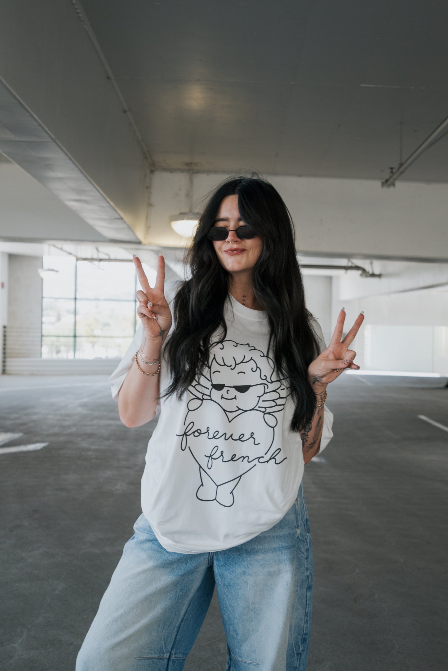 A woman with long dark hair, wearing sunglasses, jeans, and the “Adult Cotton Tee” by forever french baby featuring a white “forever friend” graphic, stands in a parking garage making peace signs and smiling—showcasing this unisex everyday cotton t-shirt.
